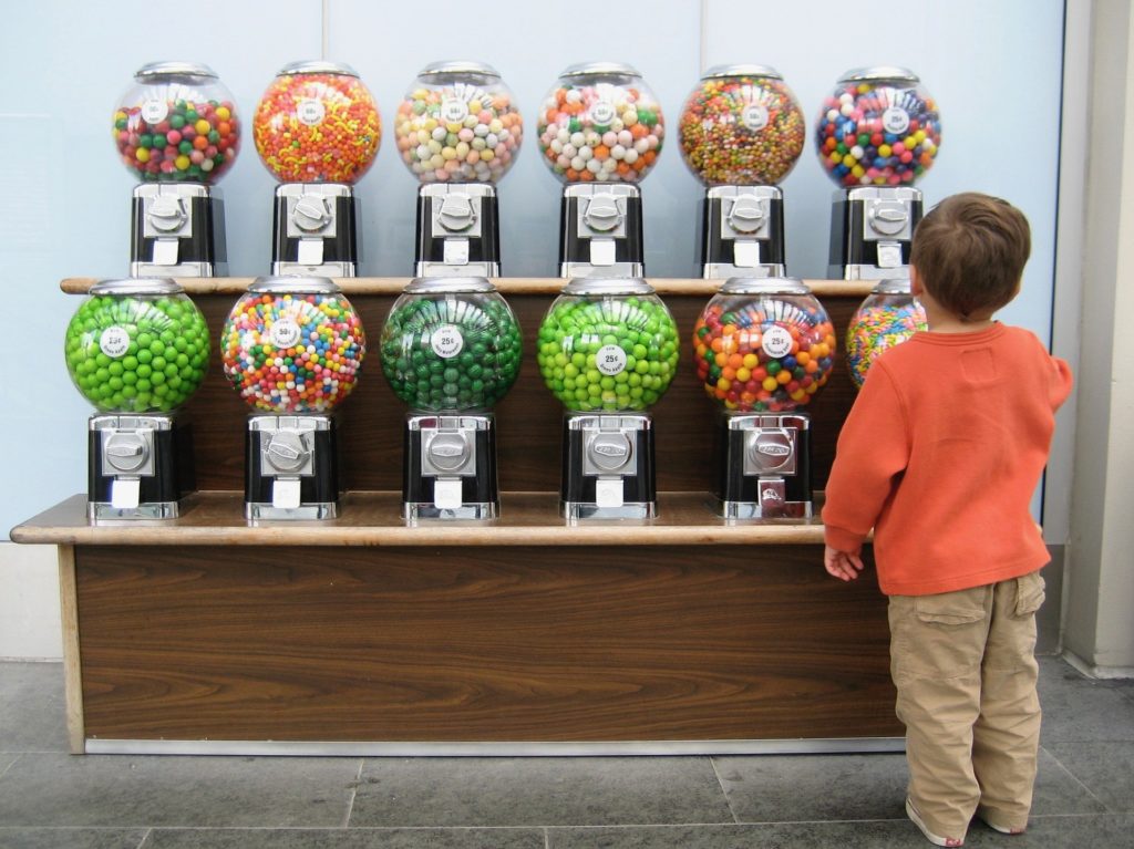 Op deze afbeelding staat een jongetje dat een juiste keuze probeert te maken tussen alle snoepautomaten met kauwgumballen.